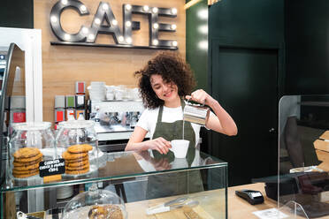 Erfreute ethnische weibliche Barista in Schürze stehen an der Theke im Café und gießen Milch in Tasse mit aromatischen Kaffee - ADSF37850