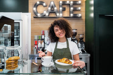 Glad ethnic female barista in apron standing at counter with cup of aromatic cappuccino and fresh croissant while looking at camera - ADSF37851