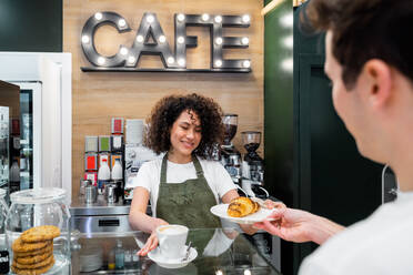 Freundliche ethnische weibliche Barista serviert eine Tasse Cappuccino und ein frisches süßes Croissant für einen Kunden, während er an der Theke eines Cafés steht - ADSF37852