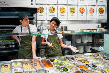 Delighted multiethnic male and female sellers in uniform and gloves standing at counter with assorted ingredients for salads in supermarket - ADSF37854