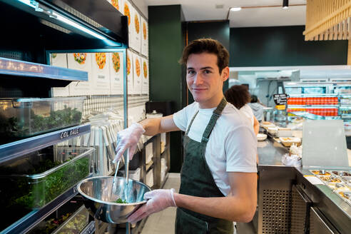 Seitenansicht eines lächelnden männlichen Verkäufers in Schürze, der grüne Kräuter in eine Schüssel gibt, während er in einer Salatbar im Supermarkt arbeitet und in die Kamera schaut - ADSF37855