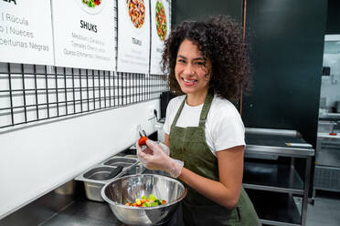 Positive ethnische Verkäuferin mit Handschuhen und Schürze, die frisches Gemüse schneidet, während sie Salat zubereitet und in einer Bar im Supermarkt arbeitet - ADSF37859