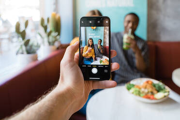 Unkenntlich gemachte männliche Person, die ein Foto von lächelnden multiethnischen Partnern mit dem Handy am Tisch mit Essen im Restaurant macht - ADSF37867