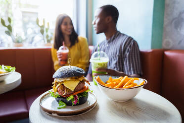 Leckerer schwarzer Burger und frittierte Kartoffelstäbchen in Schale auf Tisch gegen multirassische Partner mit Smoothies sprechen in Restaurant - ADSF37871