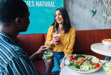Content young female with glass of smoothie talking to crop ethnic partner at table with healthy food - ADSF37873