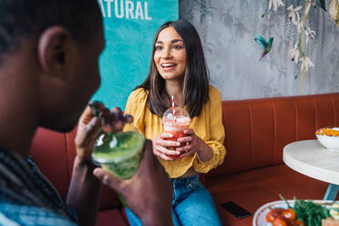 Zufriedene junge Frau mit einem Glas Smoothie im Gespräch mit einem ethnischen Partner am Tisch mit gesundem Essen - ADSF37874