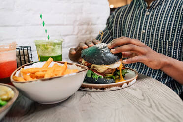 Crop anonyme ethnische männlich mit leckeren schwarzen Burger und frittierte Kartoffelstäbchen auf dem Tisch in der Nähe von Smoothie im Restaurant - ADSF37878