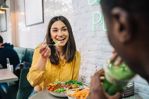 Content female enjoying tasty dish while interacting with crop unrecognizable ethnic partner drinking smoothie in restaurant - ADSF37880