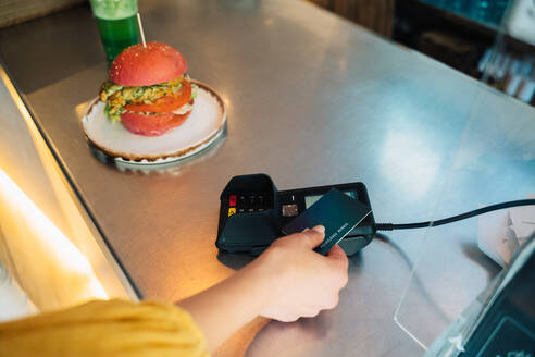 Hohe Winkel der Ernte anonyme weibliche Käufer mit Plastikkarte und Zahlungsterminal Kauf Burger an der Kasse counter - ADSF37883