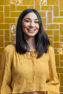 Young friendly female in yellow wear looking at camera with toothy smile against bright wall - ADSF37885