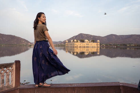 Glückliche junge asiatische Frau, die wegschaut, während sie an der Grenze in der Nähe des Sees vor dem Jal Mahal Palast und den Bergen in Jaipur, Indien, steht - ADSF37913