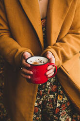 Von oben Ernte Frau in warmen braunen Mantel und geblümten Kleid hält lebhaften roten Metallbecher mit heißem Getränk beim Picknick im Herbst Park - ADSF37936