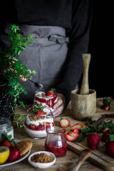 Crop person in black shirt and gray apron cooking delicious dessert with cheese and fresh ripe strawberry and cookies at table against black background - ADSF37938