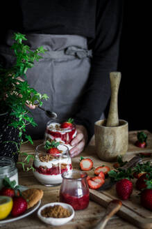 Crop Person in schwarzem Hemd und graue Schürze Kochen köstlichen Dessert mit Käse und frischen reifen Erdbeeren und Kekse am Tisch gegen schwarzen Hintergrund - ADSF37938
