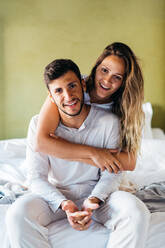 Lächelnde junge Freundin mit langen Haaren umarmt sanft glücklich Freund, während zusammen auf bequemen Bett in der Tageszeit sitzen und Blick auf die Kamera - ADSF37988