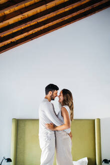 Seitenansicht eines glücklichen jungen Paares in weißer Kleidung, das sich auf dem Bett an den Händen hält und die Nasen berührt - ADSF37989