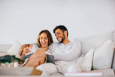 Unrasierter Ehemann in häuslicher Kleidung, der seine glückliche Frau umarmt, während er zusammen auf einer bequemen Couch sitzt und ein interessantes Buch zu Hause liest - ADSF37990