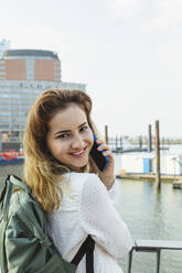 Lächelnde Frau, die an der Reling im Hamburger Hafen steht und mit ihrem Smartphone spricht, Deutschland - IHF01234