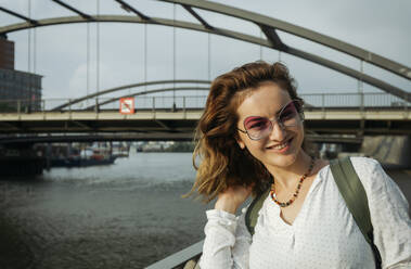 Happy woman wearing eyeglasses standing in front of bridge on sunny day, Hamburg, Germany - IHF01236