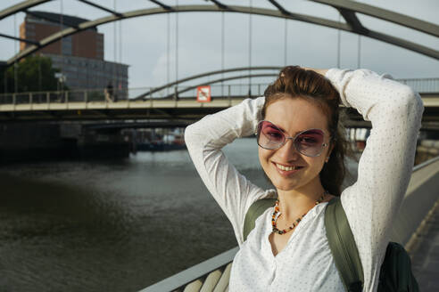 Glückliche Frau mit Händen hinter dem Kopf vor einer Brücke stehend, Hamburg, Deutschland - IHF01237