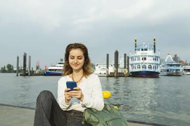 Smiling woman using smart phone sitting at Port of Hamburg, Germany - IHF01241