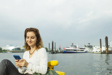 Lächelnde Frau, die ein Mobiltelefon benutzt, sitzend im Hamburger Hafen, Deutschland - IHF01243