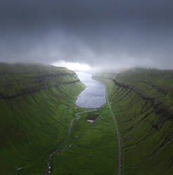 Drone view of curvy roads going on green mossy fjord near calm sea water against gray overcast sky on stormy day on Faroe Islands - ADSF37992