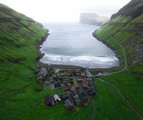 Von oben kleine Siedlung an einem grünen Fjord in der Nähe des wogenden Meeres an einem stürmischen Tag auf den Färöer Inseln - ADSF37998
