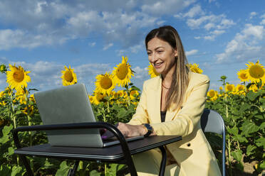 Glückliche Geschäftsfrau mit Laptop in einem Sonnenblumenfeld - OSF00909