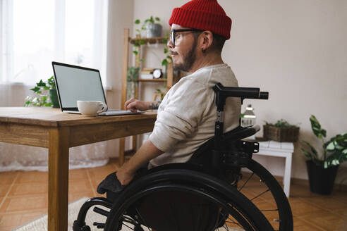 Nachdenklicher Mann im Rollstuhl mit Laptop zu Hause - EKGF00023