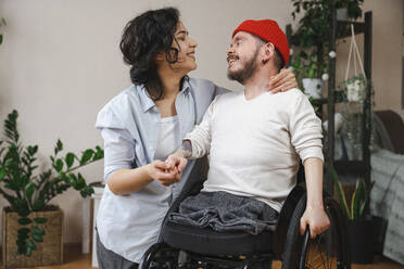 Smiling man in wheelchair holding girlfriend's hand at home - EKGF00039