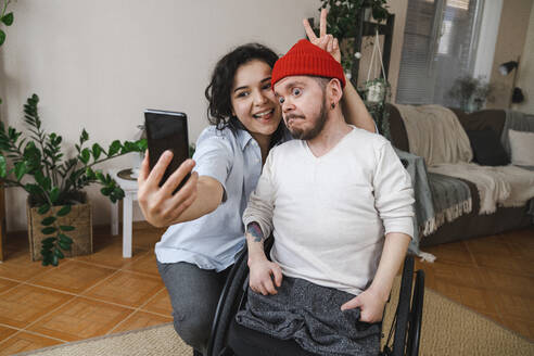 Glückliche Frau mit Mann im Rollstuhl nimmt Selfie durch Handy zu Hause - EKGF00042