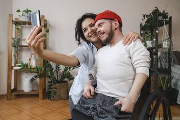 Lächelnde Frau mit Freund im Rollstuhl, die ein Selfie mit ihrem Handy zu Hause macht - EKGF00043