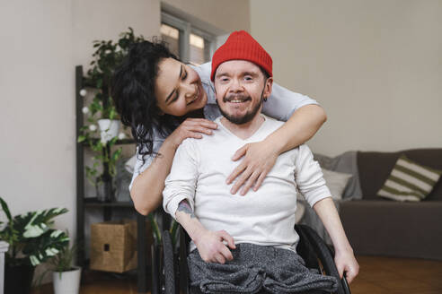 Lächelnde Frau umarmt ihren Freund im Rollstuhl zu Hause - EKGF00044