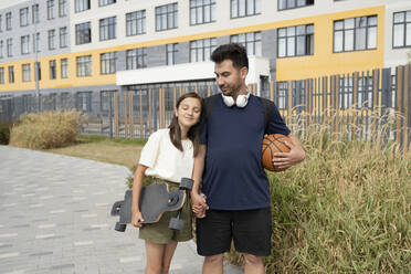 Lächelndes Mädchen mit geschlossenen Augen hält die Hand des Vaters, der einen Basketball vor einem Gebäude trägt - LESF00193