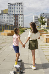 Girl holding hand of sister teaching longboard skating at footpath on sunny day - LESF00211