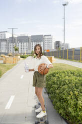 Lächelndes Mädchen mit Basketball übt Longboard-Skaten am Fußweg - LESF00212