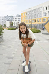 Fröhliches Mädchen mit Basketball übt Longboard-Skaten auf dem Gehweg - LESF00215