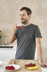 Lächelnder Mann mit Teetasse neben Früchten in der Küche stehend - VEGF05934