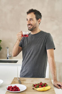 Smiling man with tea cup standing by fruits in kitchen - VEGF05934