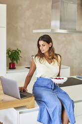 Freelancer with bowl of fruits working on laptop in kitchen - VEGF05936