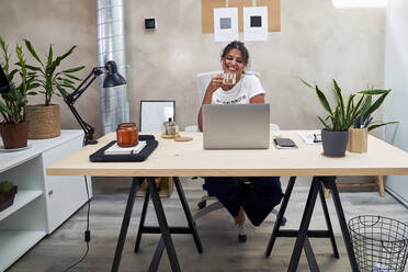 Smiling businesswoman drinking tea doing video call through laptop at home office - VEGF05949