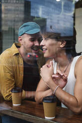 Smiling gay couple with coffee cups seen through window of cafe - ASGF02847