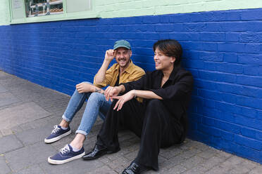 Lächelndes schwules Paar auf dem Bürgersteig vor einem Gebäude sitzend - ASGF02850