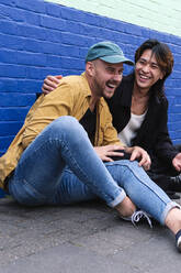 Glückliches schwules Paar sitzt auf dem Bürgersteig vor einer Mauer - ASGF02851