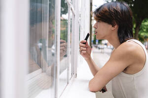 Junger schwuler Mann trägt Lippenstift auf und schaut zum Fenster - ASGF02878