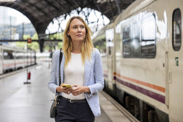Geschäftsfrau mit Smartphone, die im Bahnhof am Zug vorbeigeht - WPEF06360