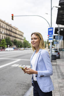 Lächelnde Geschäftsfrau mit Smartphone, die am Bürgersteig auf eine Mitfahrgelegenheit wartet - WPEF06373