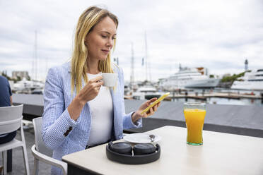 Businesswoman using smart phone holding coffee cup sitting at cafe - WPEF06378
