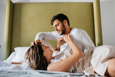 Young couple wearing similar sleepwear lying down on comfortable bed with white bedding and looking at each other - ADSF38015